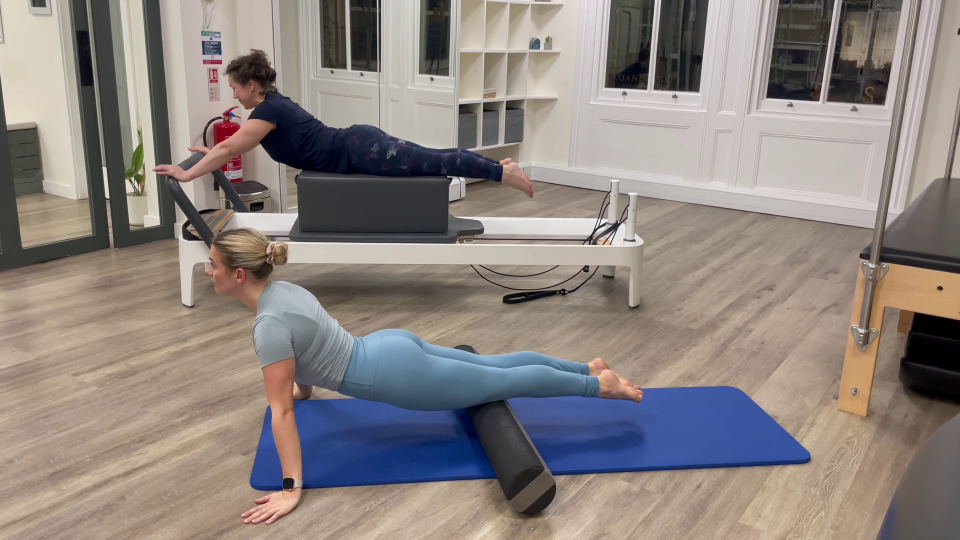Two women performing Pilates exercises; one on a reformer, the other using a foam roller.