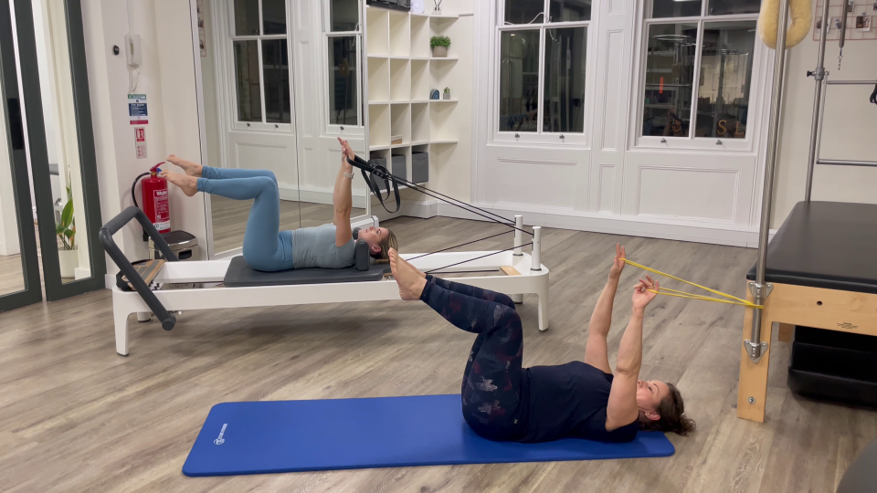 Two women performing Pilates exercises using resistance bands and a reformer machine.