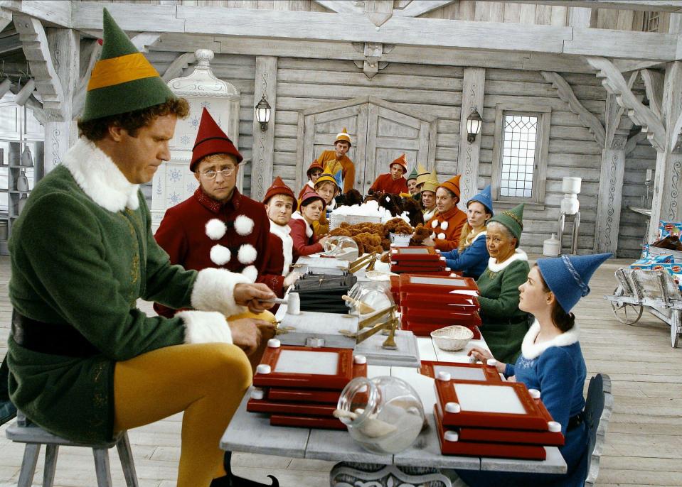 A still from the movie Elf, starring Will Ferrell and Zooey Deschanel. Ferrell is dressed as an elf and is standing in front of an elf door in a snowy forest. Deschanel is standing next to him, dressed as a human. Both are looking at the camera.
