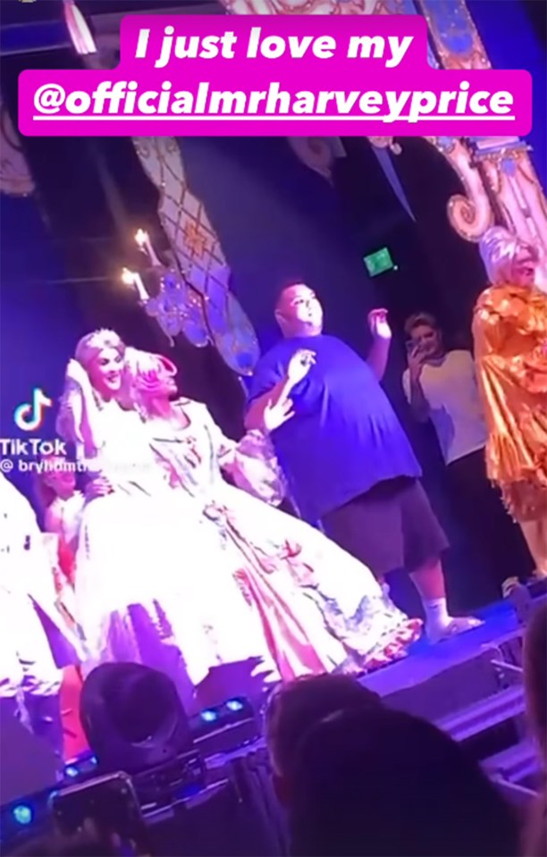 A man in a blue shirt stands with two people in elaborate dresses on stage.