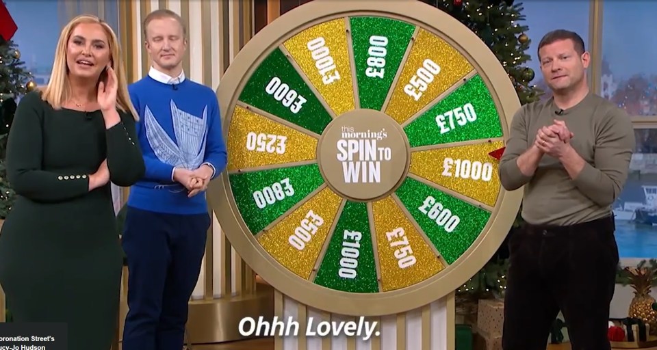 Three people stand beside a prize wheel on a game show.