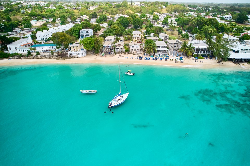 Aerial view of Alleynes Bay, Barbados, showing a beach, turquoise water, boats, and buildings.