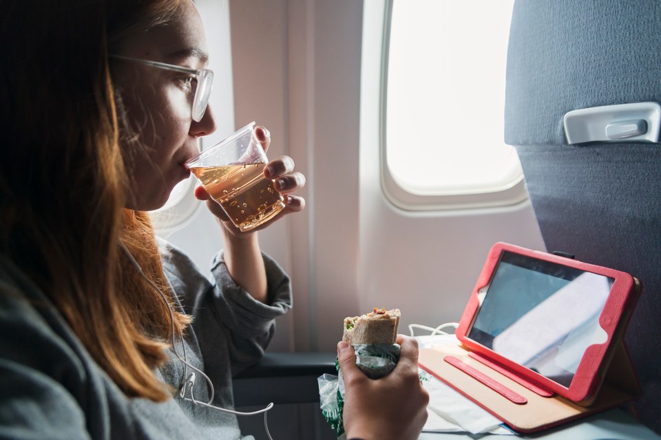 Woman on airplane watching movie, eating sandwich, and drinking ginger ale.