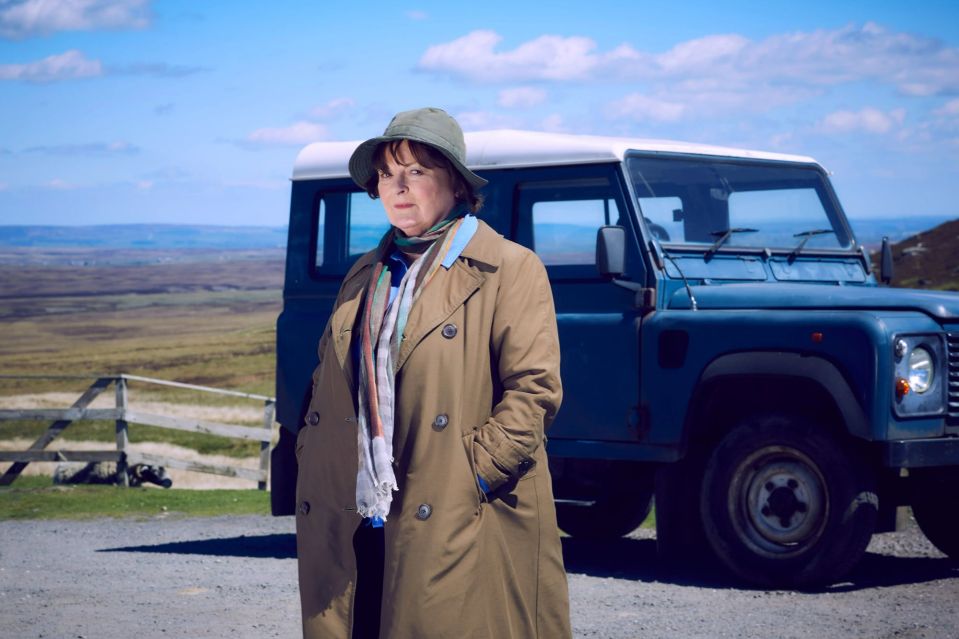 A woman in a trench coat stands beside a Land Rover in a rural setting.