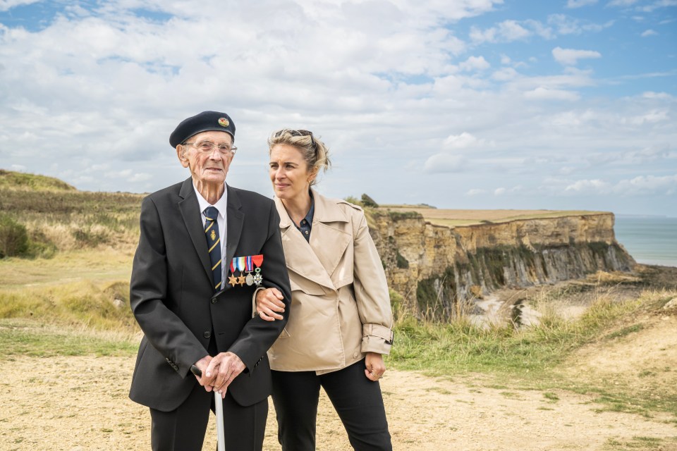 A woman and her grandfather stand together on a cliff overlooking the ocean.