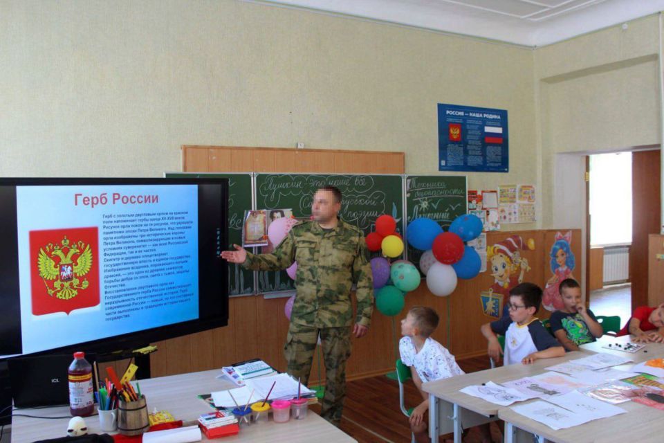 A soldier in camouflage giving a presentation about the Russian coat of arms to a group of Ukrainian children.