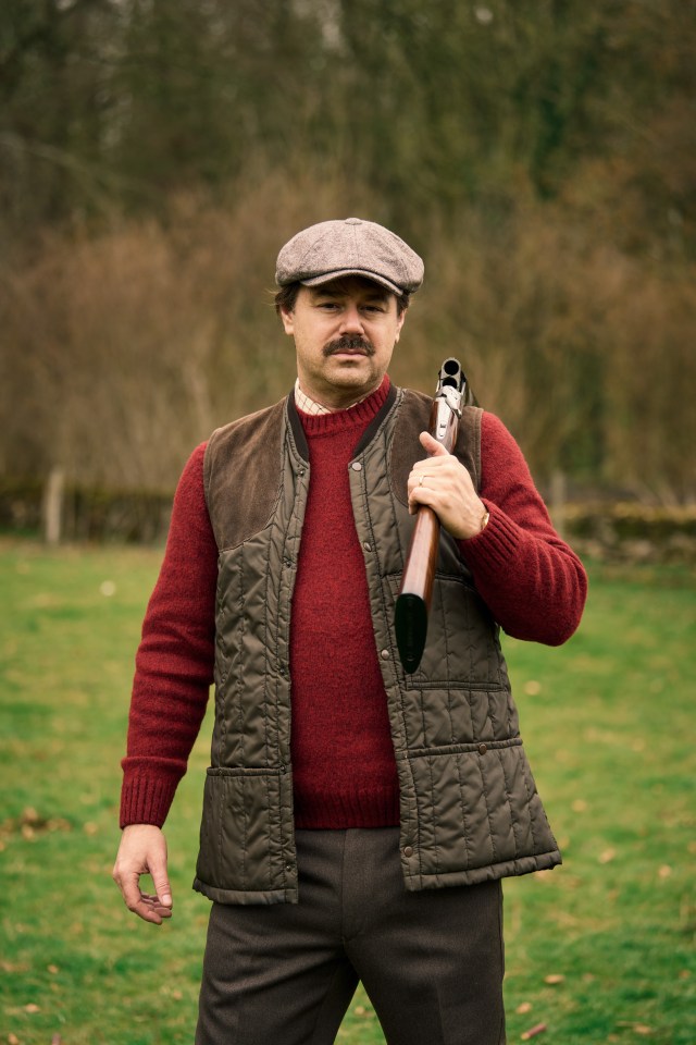 Man in tweed cap and shooting vest carries a shotgun.