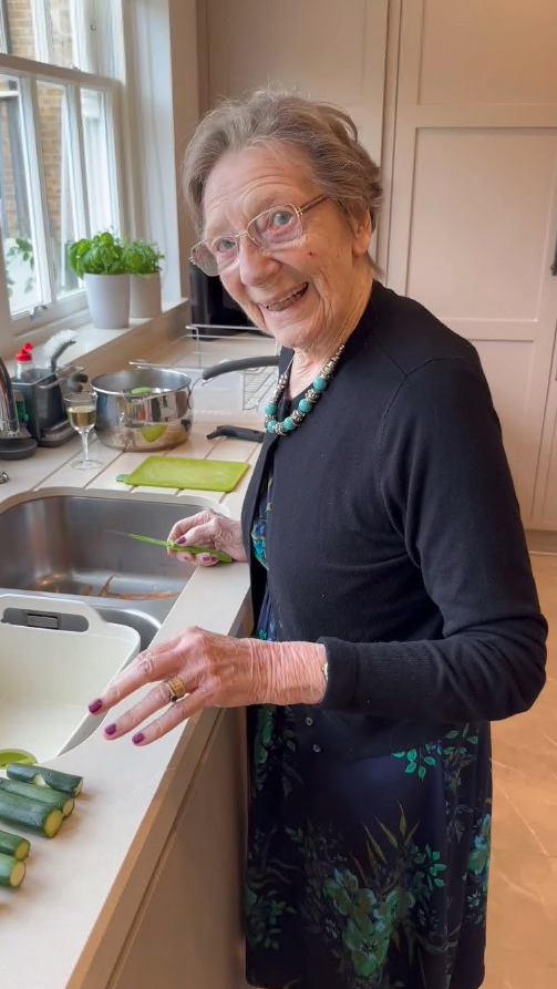 A smiling older woman prepares zucchini in her kitchen.