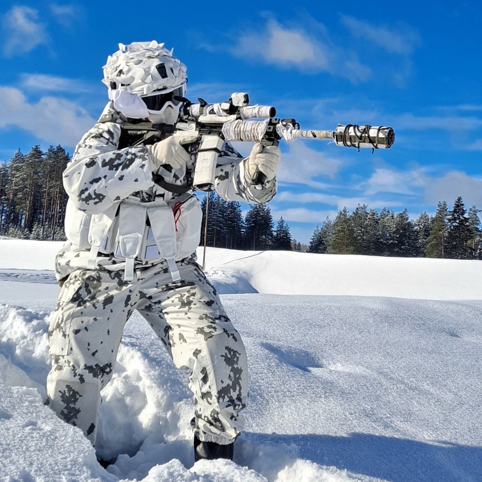 Finnish soldier in winter camouflage aiming a rifle in snowy landscape.
