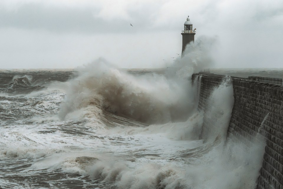 Large waves crashing against a lighthouse and seawall.