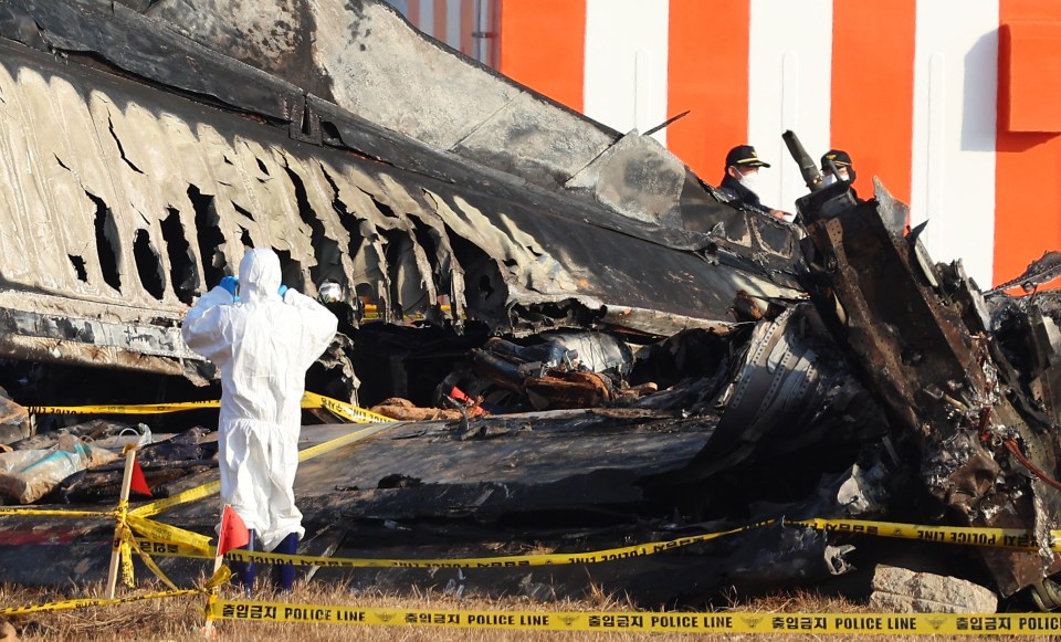 Debris from a plane crash at an airport, with investigators in protective suits at the scene.