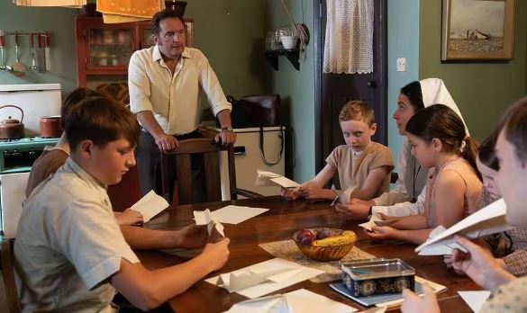 A man stands behind a table of children making paper airplanes.