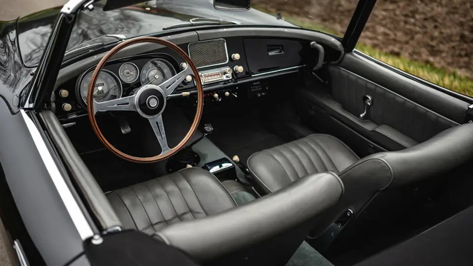 Interior view of a black 1958 BMW 507 Roadster, showing grey leather seats, wooden steering wheel, and dashboard details.
