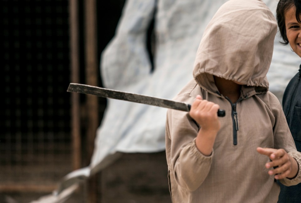 A hooded child holds a machete.