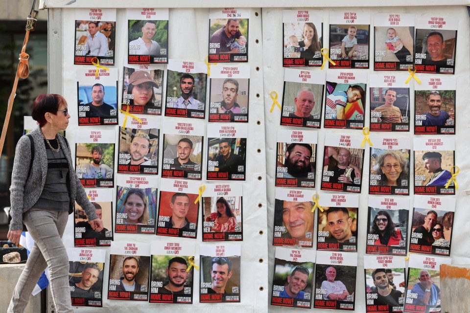 Photo of a woman walking past a board displaying photos of Israeli hostages with the text "Bring Him/Her Home Now!" in Hebrew and English.
