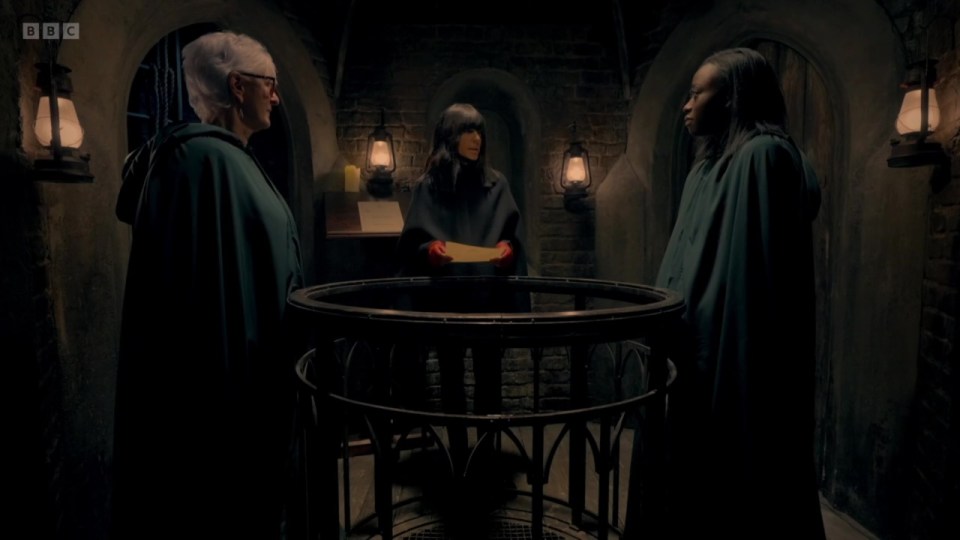 Three women in cloaks stand around a circular railing, one holding a piece of paper.