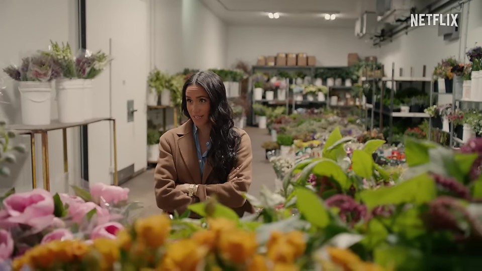 Meghan Markle standing in a flower warehouse with arms crossed.