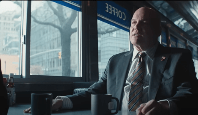 A man in a suit sits in a diner, looking thoughtfully out the window.