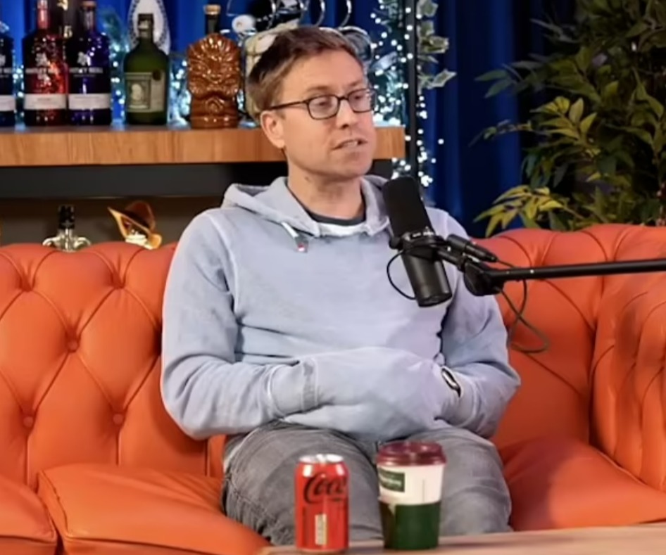 Man in light blue hoodie sits on an orange couch during a podcast recording.