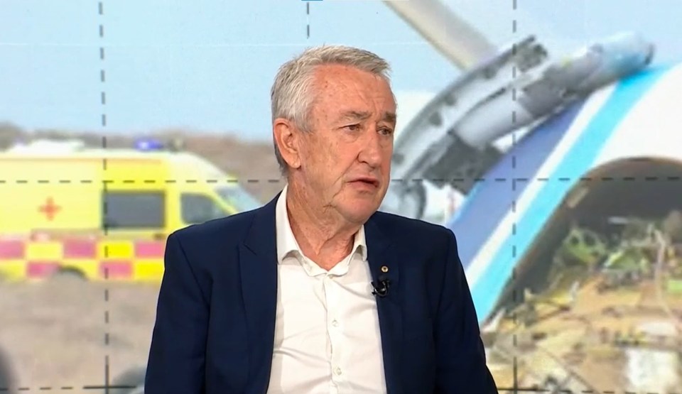Man in suit being interviewed, with a plane crash in the background.