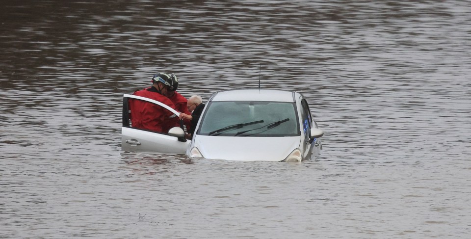 Firefighters rescuing a man from a car submerged in floodwater.