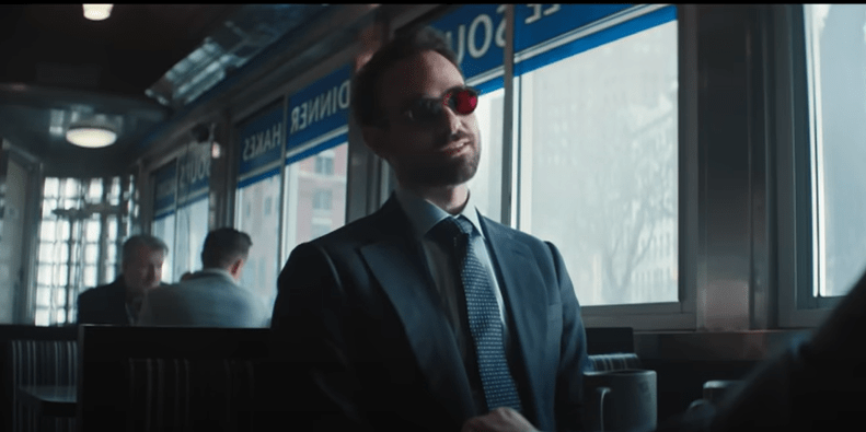 Matt Murdock, Daredevil, in a suit and red glasses, sits in a diner.