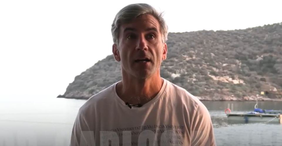A man speaks in front of a body of water and a hillside.