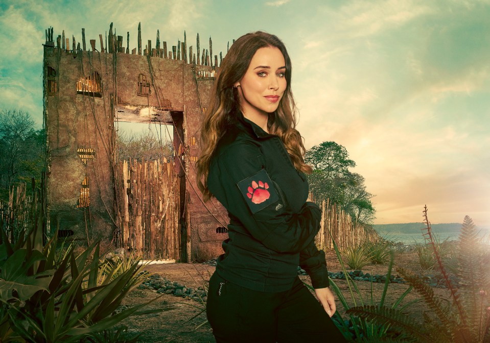 Una Healy in a black jacket with a paw print patch, standing before a rustic wooden gate.