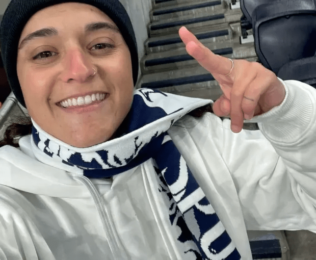 Smiling woman wearing a white hoodie and a navy and white scarf, giving a peace sign.