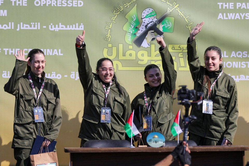 Four women in military-style uniforms stand at a table, giving thumbs up.