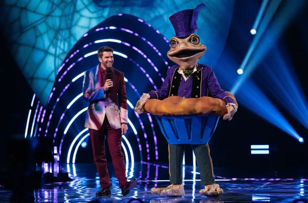 A man in a colorful suit stands next to a contestant dressed as a frog in a pie costume on a stage.