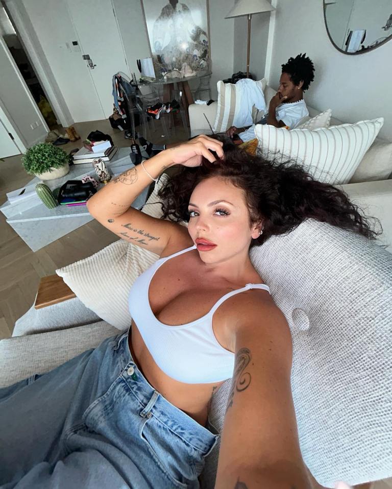 Jesy Nelson in a white bikini top and jeans, lounging on a couch with Zion Foster in the background.