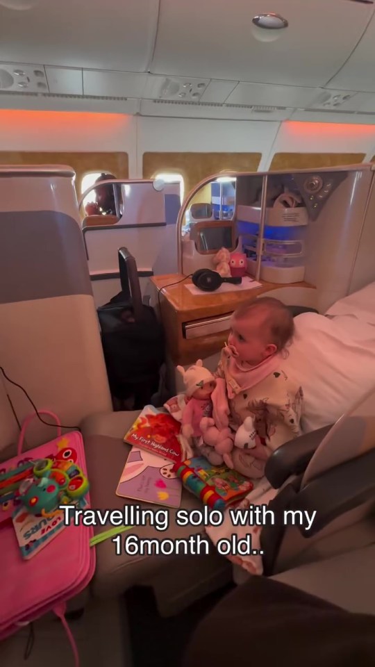 A baby sits in a first-class airplane seat surrounded by toys and books.