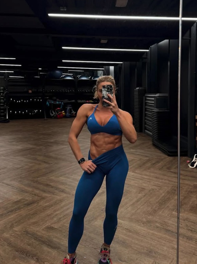 Gabby Allen in a gym, showing off her physique in blue workout clothes.