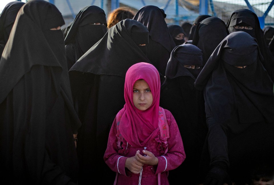 A young girl in a pink headscarf stands among women in black niqabs at the al-Hol camp.