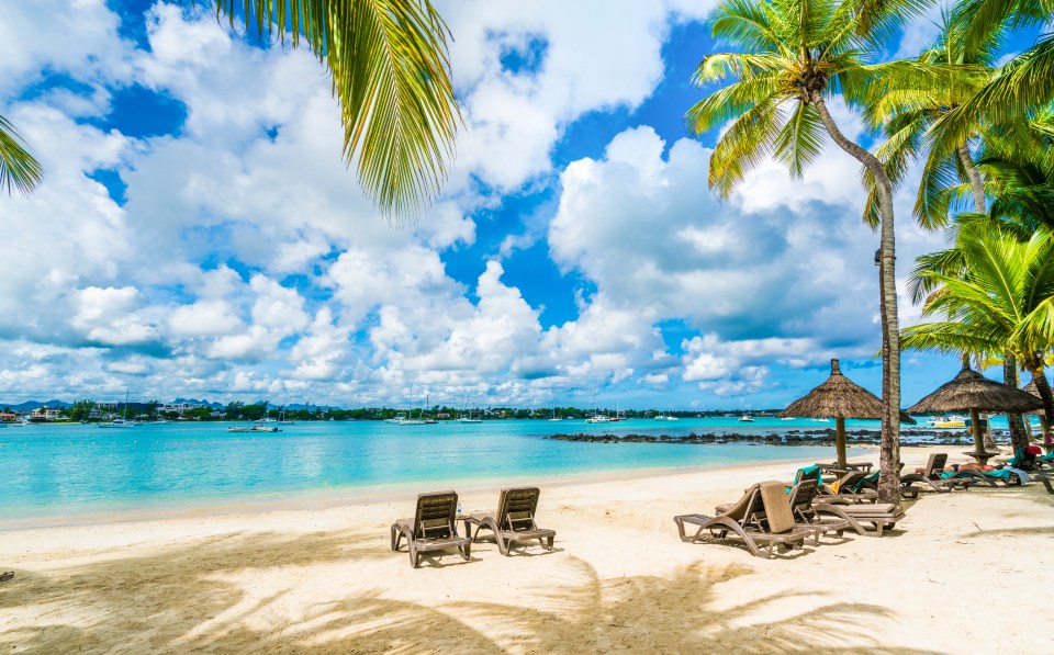 Beach scene in Grand Baie, Mauritius, with lounge chairs under thatched umbrellas and palm trees.