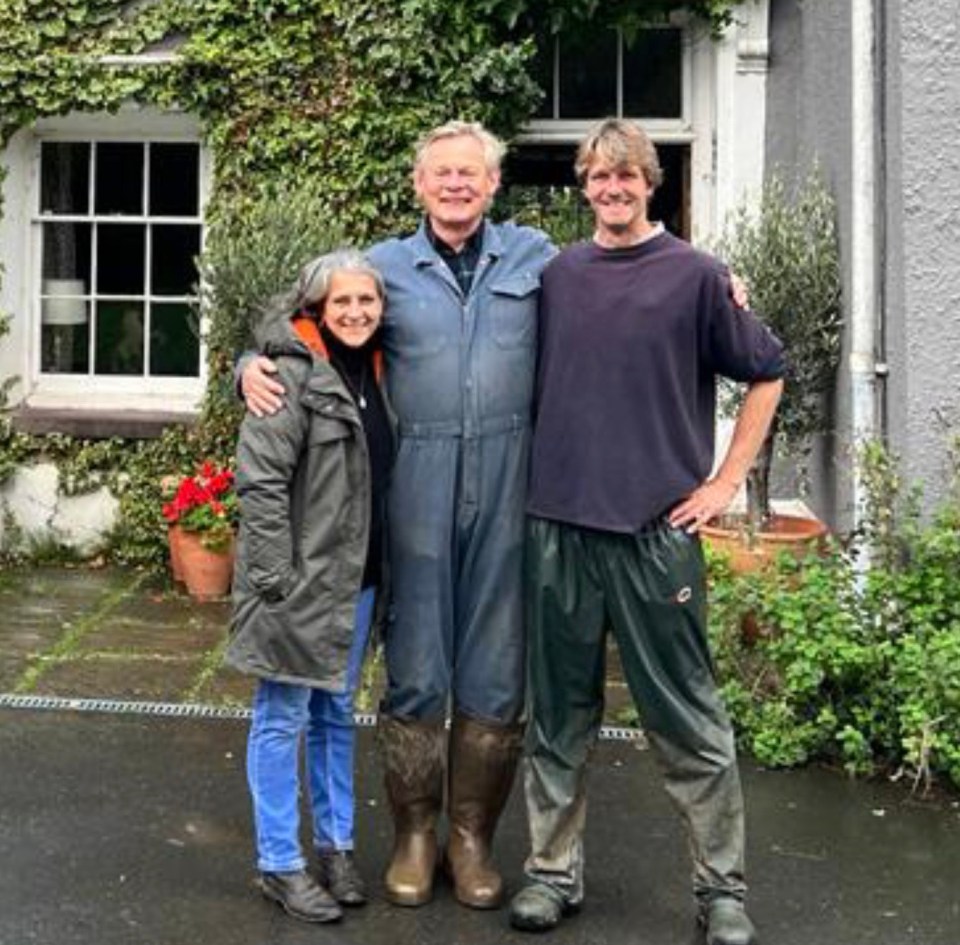 A couple and a man stand outside a farmhouse.