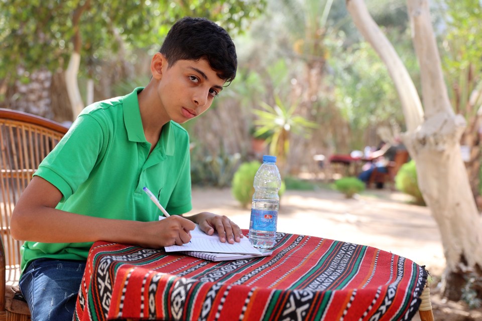 A boy sits at a table outdoors writing in a notebook.
