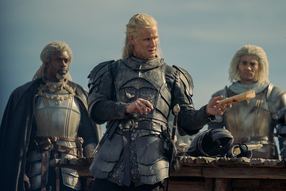 A man in dark armor reads a scroll to two other armored men.