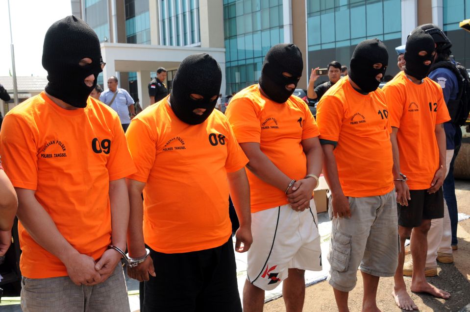 Indonesian police officers in orange shirts and balaclavas display suspects arrested for producing and selling illegal alcohol.