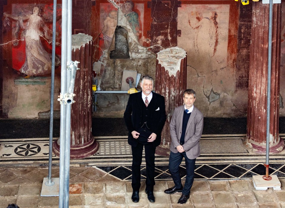 Two men standing in front of a partially excavated Roman fresco.