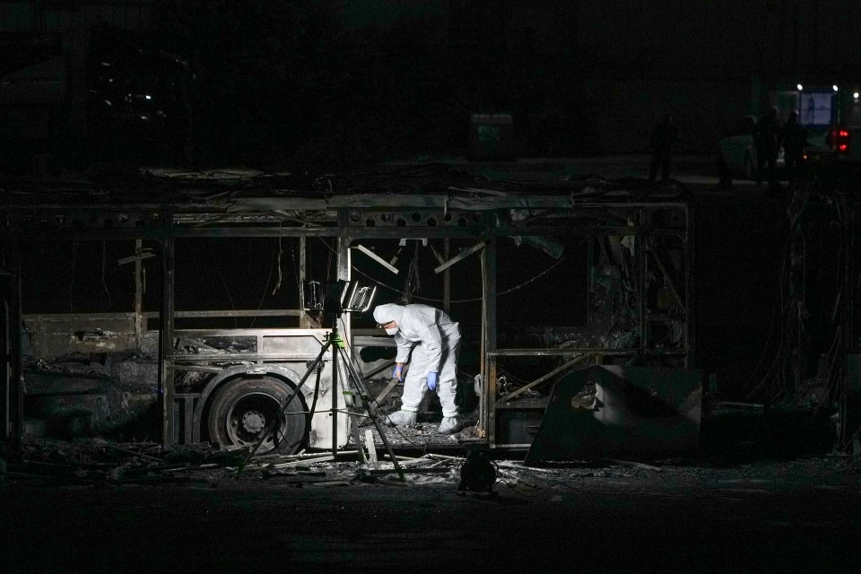 A forensic investigator examines a burned-out bus at night.