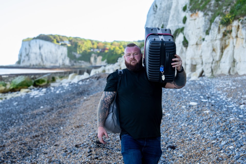 Man carrying a suitcase on a pebble beach with white cliffs in the background.