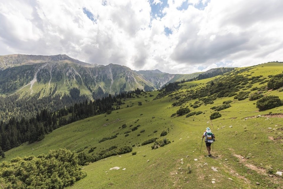 A hiker walks a trail in a mountainous, grassy landscape.