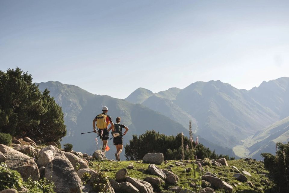 Two trail runners with backpacks run along a rocky mountain path.