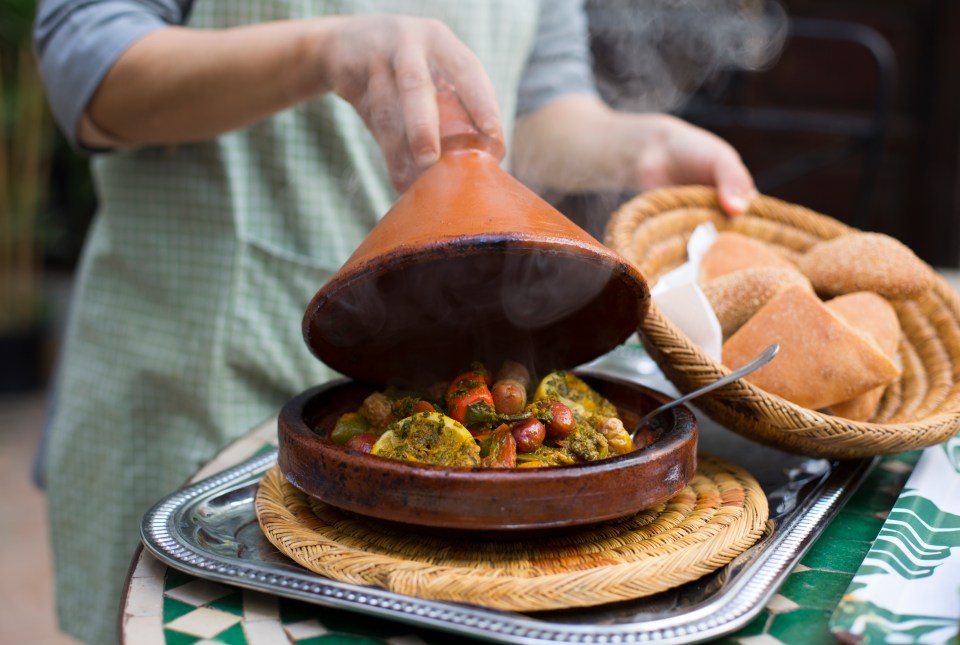 A woman lifts the lid off a tagine of steaming Moroccan stew.