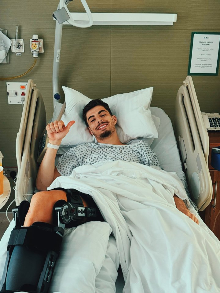 Man in hospital bed with knee brace, giving thumbs up.