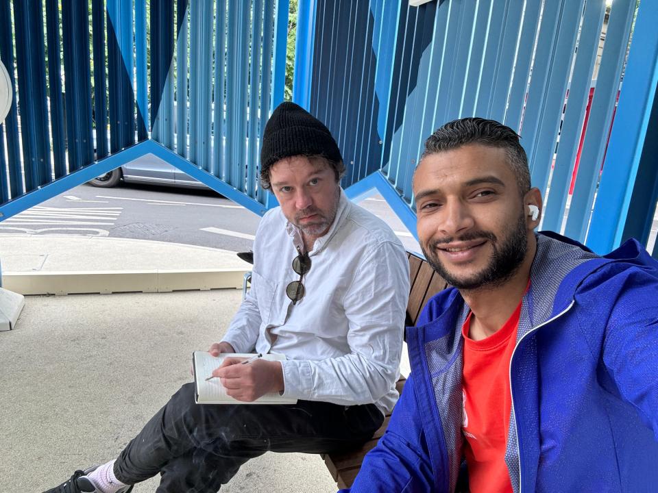 Two men sitting on a bench, one writing in a notebook.