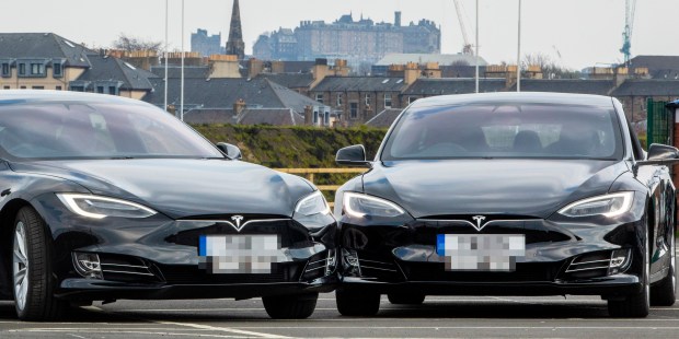 Two black Tesla cars parked in a lot with a city in the background.