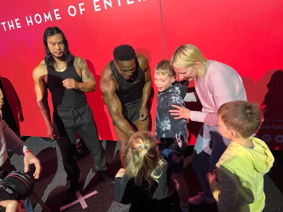 Children meeting muscular men in front of a red wall.